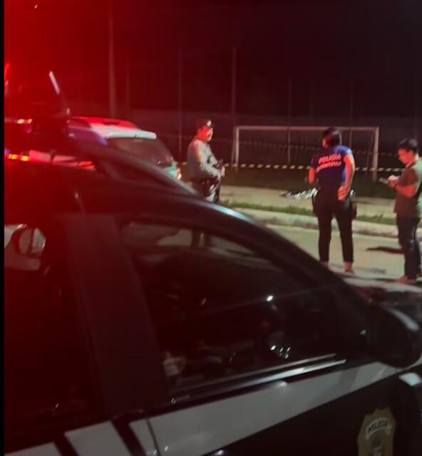 foto mostra, em primeiro plano, carro preto da polícia civil. ao fundo, policiais conversam de frente a cena do crime, cercada para perícia.